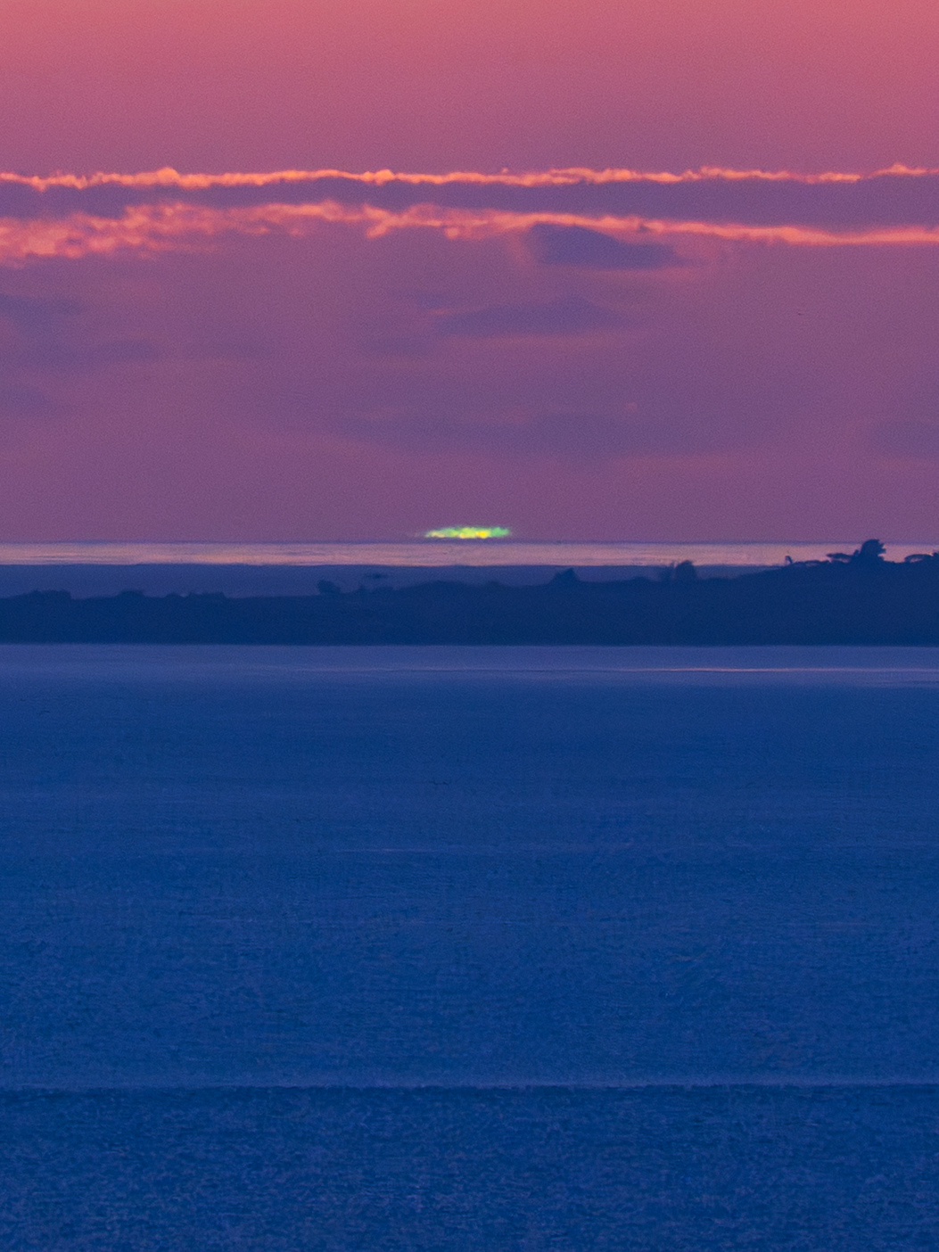 夕暮れの海の向こう、水平線近くで太陽が沈む瞬間に緑色の光が見えている。空は紫がかったピンク色に染まり、上部にはオレンジ色に縁取られた雲が横に伸びている。手前には暗い青色の海、水平線付近には島影があり、そのすぐ上に小さく鮮やかなグリーンフラッシュが輝いている。
