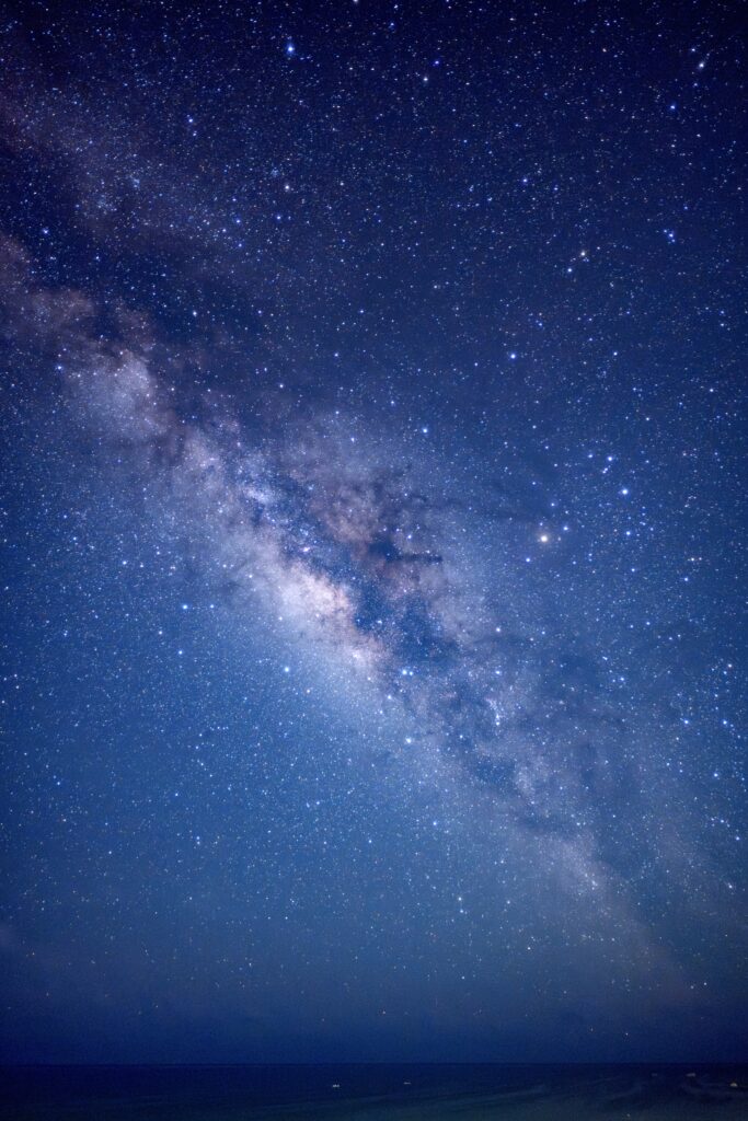 石垣島の夜空を縦構図で撮影した写真。画面下には暗い海と水平線があり、その上に夜空が広がっている。左下から中央にかけて、天の川の明るく淡い光の帯が斜めに立ち上がり、天の川の中には暗黒帯が見える。