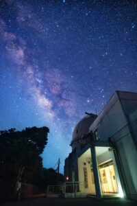 石垣島天文台の建物とドームの上に、夏の天の川が斜めに大きく昇っている夜空の写真。右下には明かりのついた天文台の建物、左下には木々のシルエットが写っている。空一面に無数の星が広がり、天の川の淡い帯が夜空に浮かび上がっている。